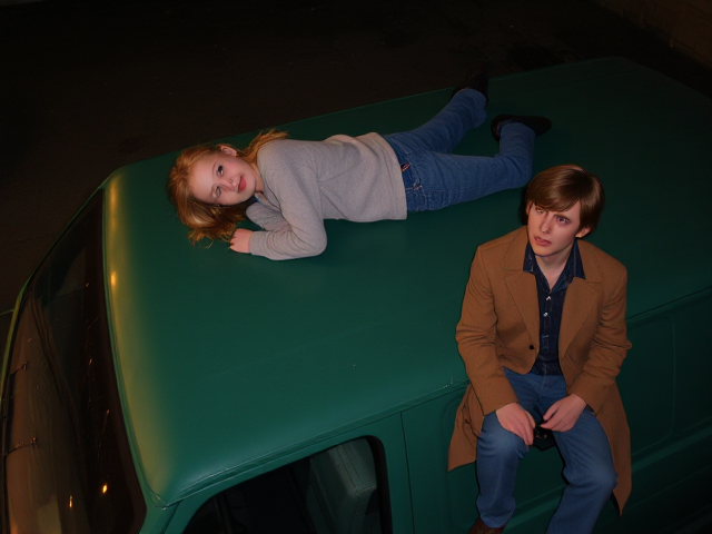 A petite, 13 year old girl with strawberry blonde hair, wearing a pullover and  jeans, laying on the roof of a green, 1987 Chevy van. She is on her back, staring up at the night sky, stargazing. Besides her sits a modern day vampire in his twenties, with blonde hair and wearing a brown trenchcoat and jeans. He looks down, uneasy.