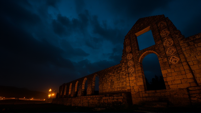 Créer une bannière, avec une ruine en dégradé ornée de motifs complexes et anciens, atmosphère brumeuse et mystique, de nuit avec un ciel étoilé, des torches enflammées illuminent les alentours avec une lumière dansante, un corbeau regardant fixement vers l'horizon, des ombres mouvantes au sol ajoutent au mystère