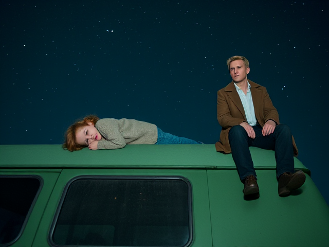 A petite 12-year-old girl with strawberry blonde hair, wearing a cozy pullover and jeans, is lying on the roof of a weathered green 1987 Chevy van. She is on her back, gazing up at the night sky filled with stars. Next to her on the roof of the van, sits a modern-day, pale skinned vampire in his late twenties, with striking blonde hair, dressed in a brown trench coat and jeans. He appears uneasy as he looks downward