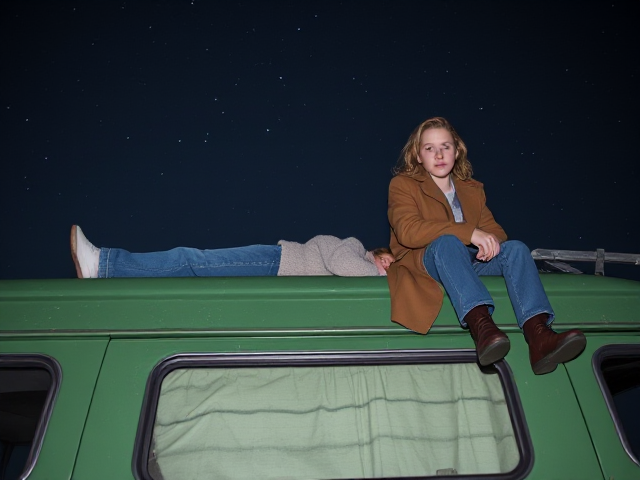 A petite 12-year-old girl with strawberry blonde hair, wearing a cozy pullover and jeans, is lying on the roof of a weathered green 1987 Chevy van. She is on her back, gazing up at the night sky filled with stars. Next to her on the roof of the van, sits a modern-day, pale skinned vampire in his late twenties, with striking blonde hair, dressed in a brown trench coat and jeans. He appears uneasy as he looks downward