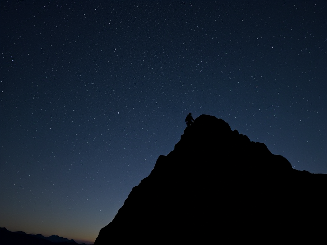 A lone mountain climber, silhouetted against a vast, starry night sky. The climber is ascending a steep, rocky mountain with determination and focus, their figure small but resolute. The landscape is serene and quiet, with the only sound imagined being the soft wind. The starry sky above conveys the idea of limitless potential. The scene is set in a peaceful, tranquil environment, emphasizing the solitude and silent effort it takes to achieve greatness, with no external recognition or distractions.