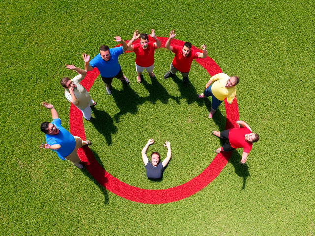 A bright, sunny day on a green grassy field. In the center, there is a bold red circle drawn on the grass. Inside the circle, there are 9 people standing or kneeling, spread evenly. They are wearing colorful, simple shirts: 3 in blue, 3 in red, and 3 in yellow. All the people are looking surprised, with their mouths open and arms raised in shock or excitement. One person is lying down inside the circle, also looking shocked. The camera angle should be slightly from above, focusing on the people in the red circle in the middle of the grassy field. The expressions of the people should be clear and normal, without any injuries or distortions. The lighting should be natural and bright, as if it’s a sunny day.