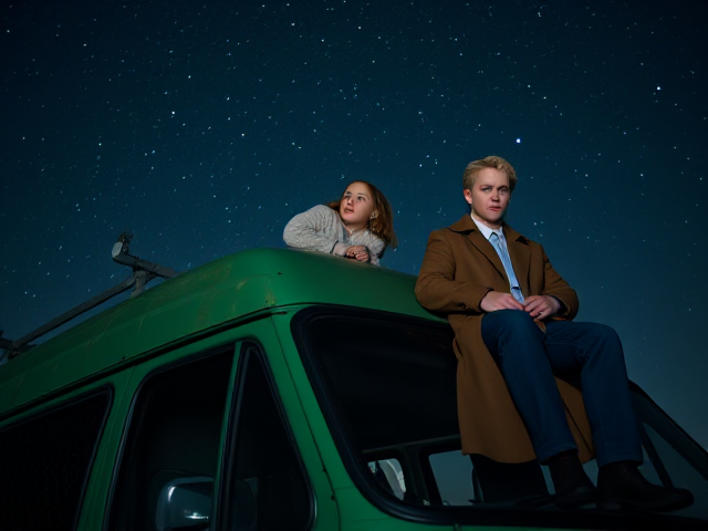 A petite 12-year-old girl with strawberry blonde hair, wearing a cozy pullover and jeans, is lying on the roof of a weathered green 1987 Chevy van. She is on her back, gazing up at the night sky filled with stars. Next to her on the roof of the van, sits a modern-day, pale skinned vampire in his late twenties, with striking blonde hair, dressed in a brown trench coat and jeans. He appears uneasy as he looks downward