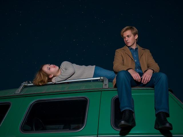 A petite 12-year-old girl with strawberry blonde hair, wearing a cozy pullover and jeans, is lying on the roof of a weathered green 1987 Chevy van. She is on her back, gazing up at the night sky filled with stars. Next to her on the roof of the van, sits a modern-day vampire in his twenties, with striking blonde hair, dressed in a brown trench coat and jeans. He appears uneasy as he looks downward
