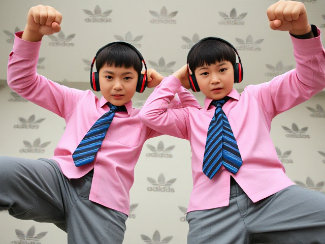 Close-up view of two fierce Japanese kids standing confidently. They have short haircuts and are stomping with one leg raised, arms raised in a dominant pose. They're wearing pink collared long-sleeve shirts with large blue striped ties, headphones on their ears, and smartwatches on their wrists. The background features Adidas logos as wallpaper. The perspective is from a low angle, emphasizing their conquering stance. Their faces display angry determination with frowned eyebrows, pursed lips, and clenched fists