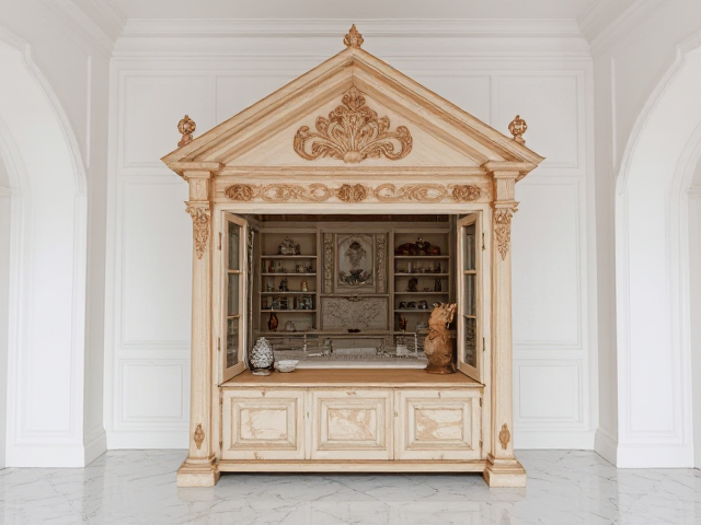 Une petite malle ancienne ouverte en bois avec des decorations style sculture en bois dans une piece entièrement blanche avec moulure au murs et sol marbre