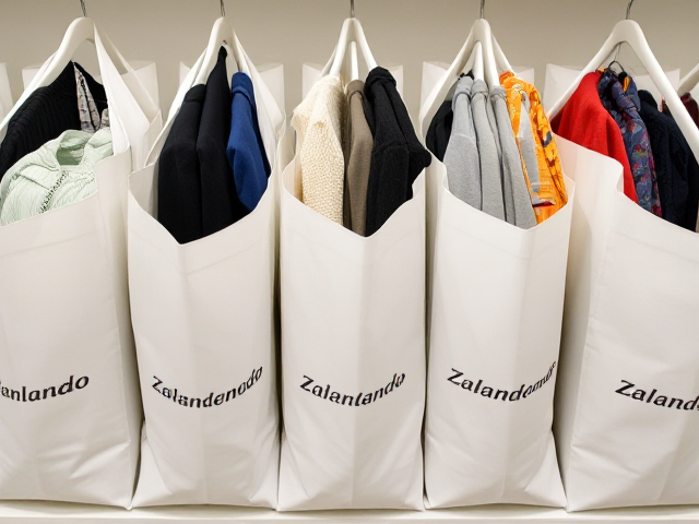 10 Einkaufstaschen, die alle gleich ausschauen. Alle weiss mit Zalando logo darauf. alle stehen geordnet nebeneinander. man sieht, dass kleider darin sind.