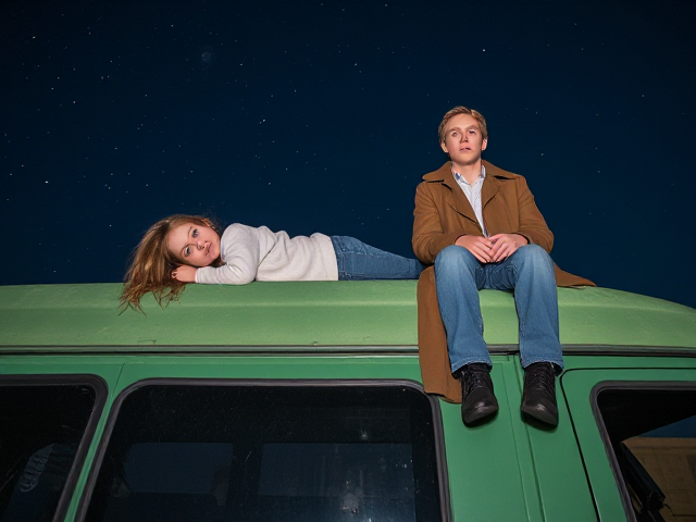 A petite 12-year-old girl with strawberry blonde hair, wearing a cozy pullover and jeans, is lying on the roof of a weathered green 1987 Chevy van. She is on her back, gazing up at the night sky filled with stars. Next to her on the roof of the van, sits a modern-day, pale skinned vampire in his late twenties, with striking blonde hair, dressed in a brown trench coat and jeans. He appears uneasy as he looks downward